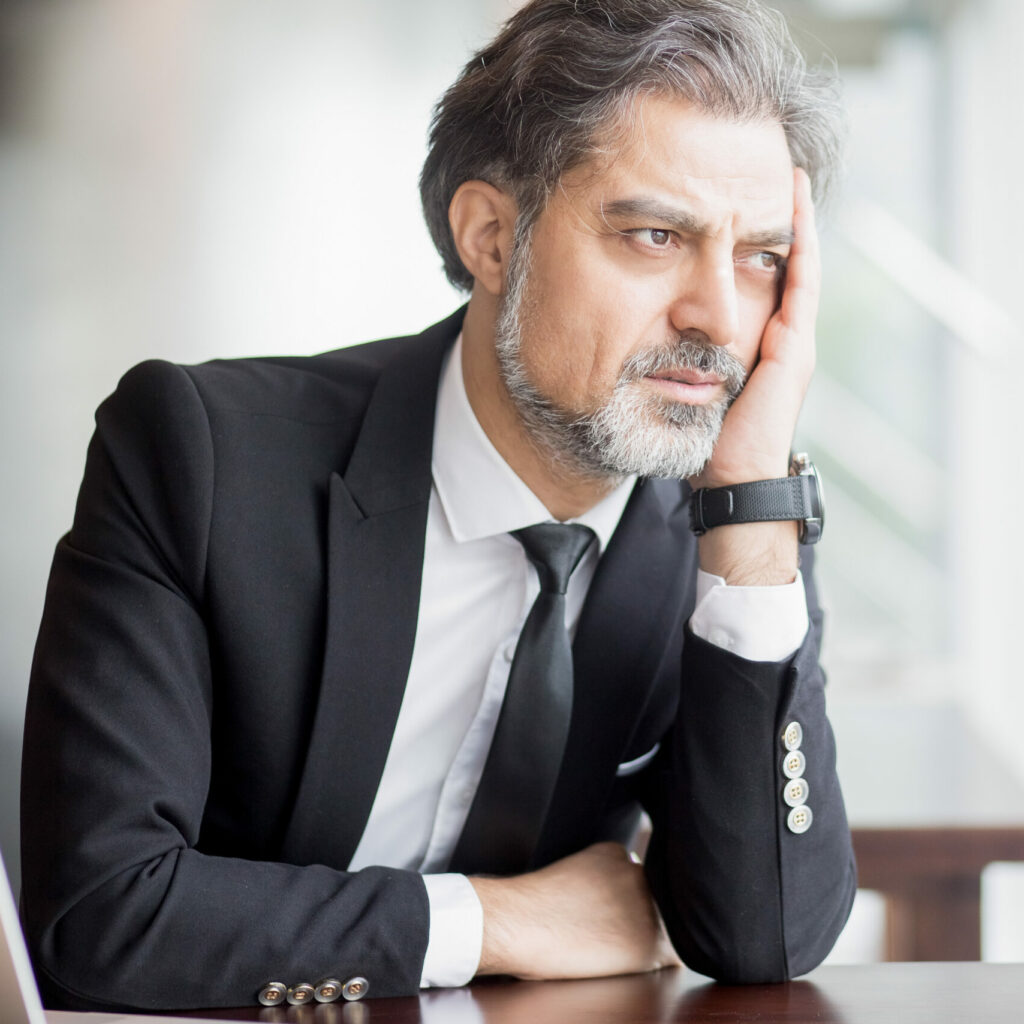 Closeup portrait of thoughtful middle-aged handsome business man looking through window, leaning head on hand and sitting at office desk