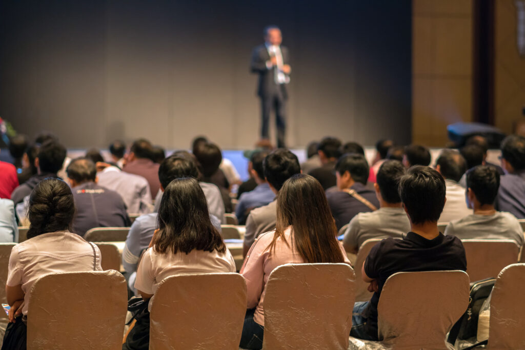 Rear side of Audiences sitting and listening the speackers on the stage in low light conference hall, event and seminar concept